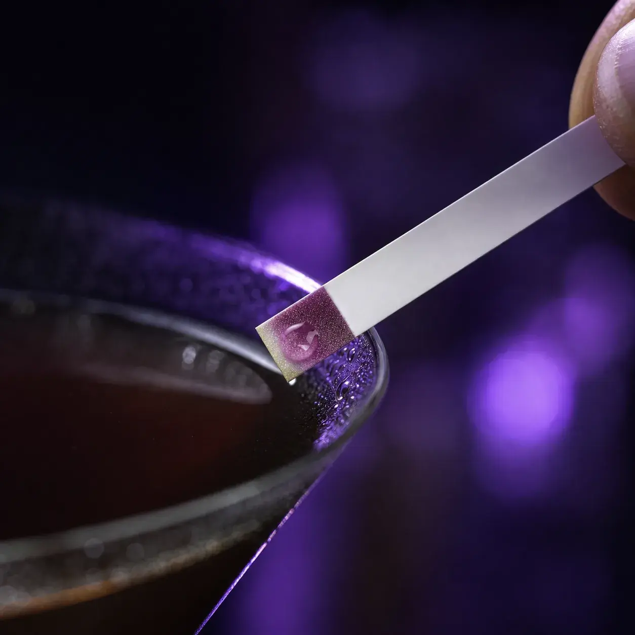 A macro shot of a drink test strip being dipped at the rim of a dark cocktail glass